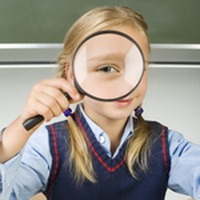 Little girl with magnifying glass in hand. Sitting at desk in front of blackboard. Magnifying her's eye. Looking at camera. Front view
